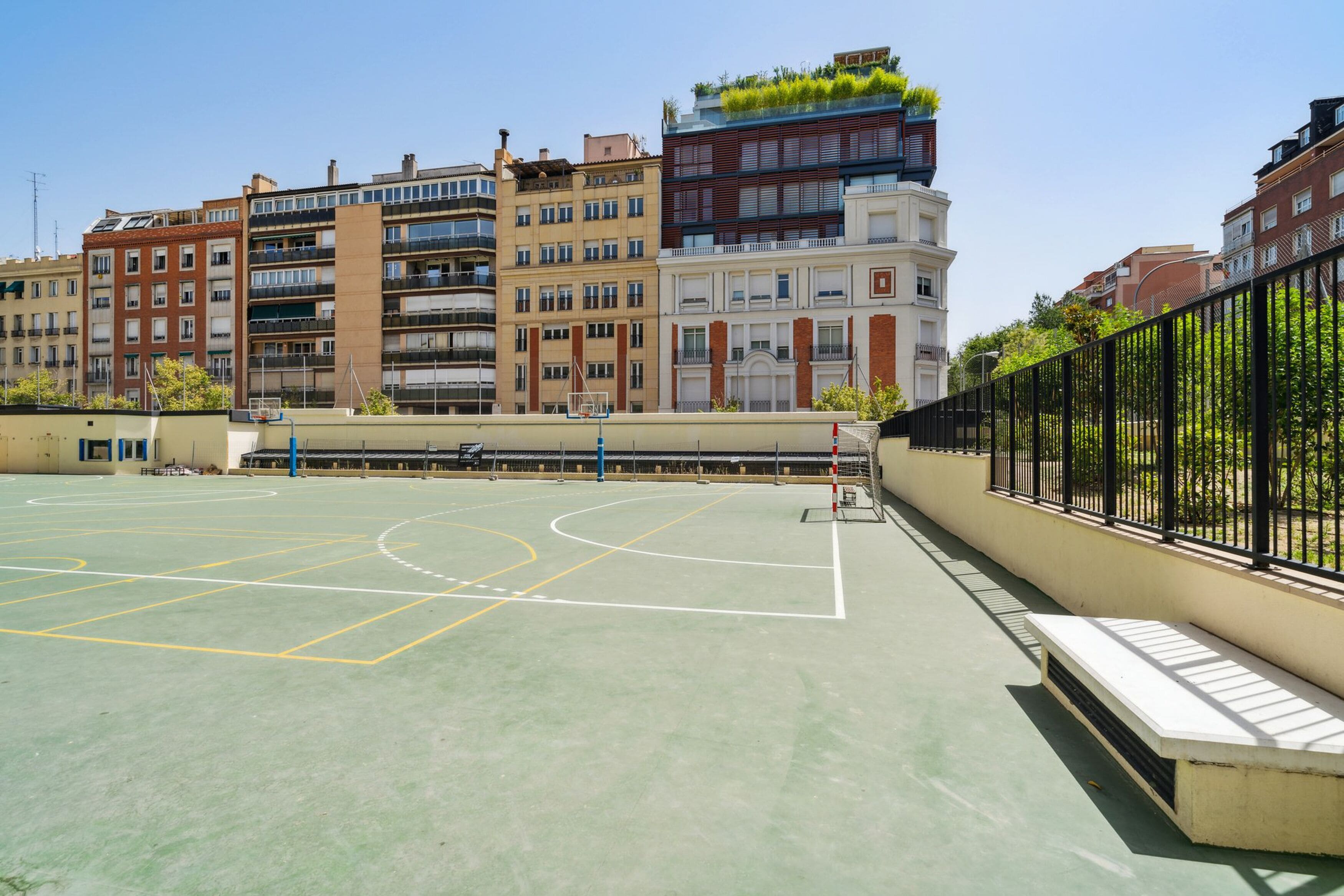 Una cancha de baloncesto al aire libre con vista despejada a edificios circundantes, incluyendo un edificio con jardín en la azotea.