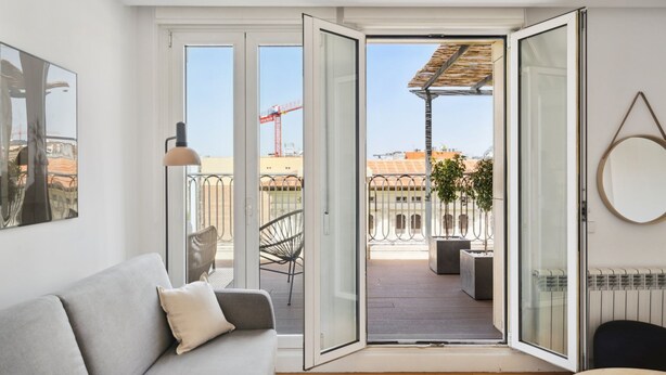A modern living room with a grey sofa, a floor lamp, and a large sliding glass door leading to an outdoor balcony with a view of the cityscape and construction cranes. - Limehome Madrid Calle de Nuñez de Balboa (Madrid)