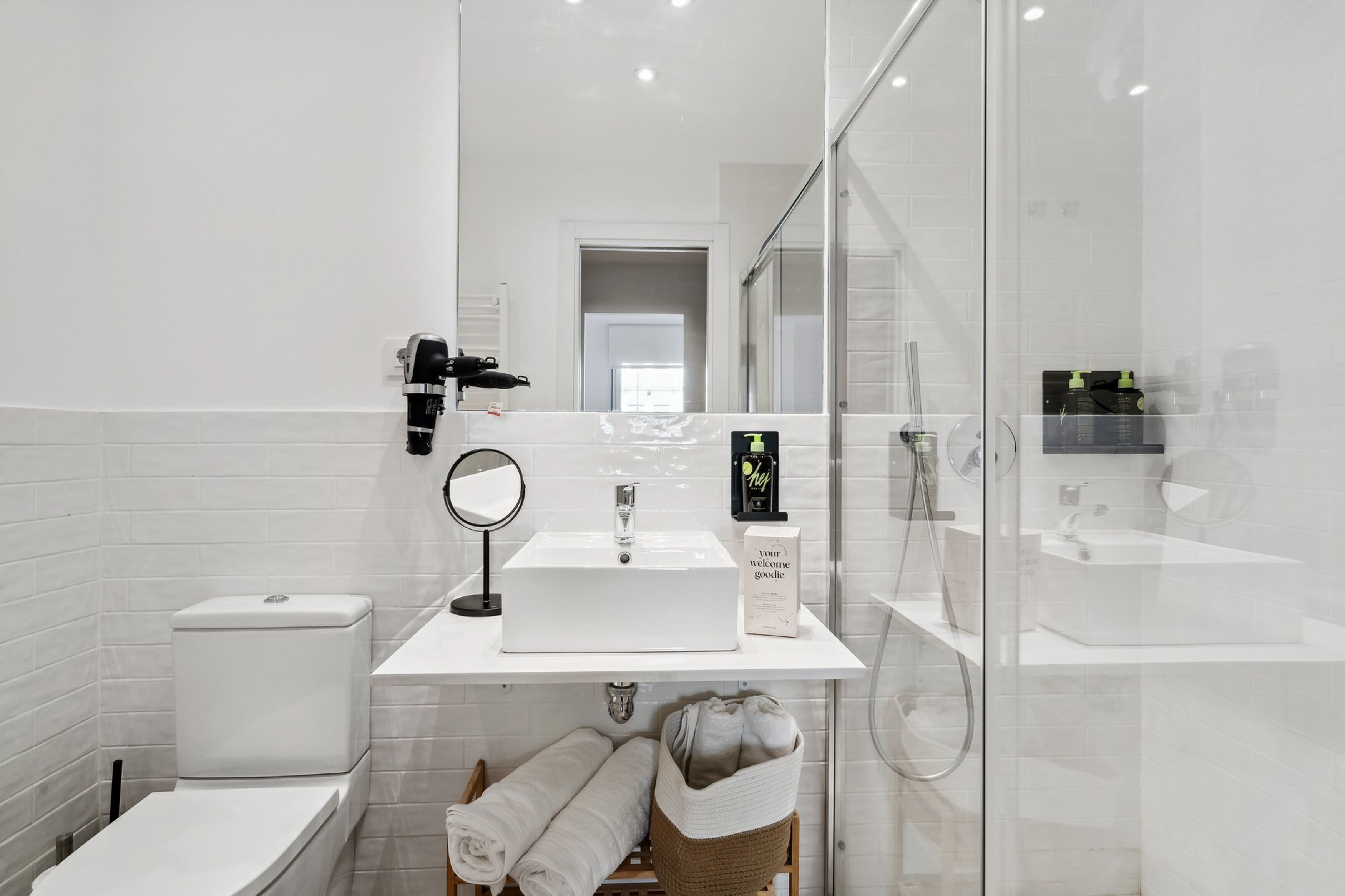 Un baño moderno con lavamanos, espejo y retrete blancos. Cuenta con un box de ducha de vidrio y un perchero con toallas.