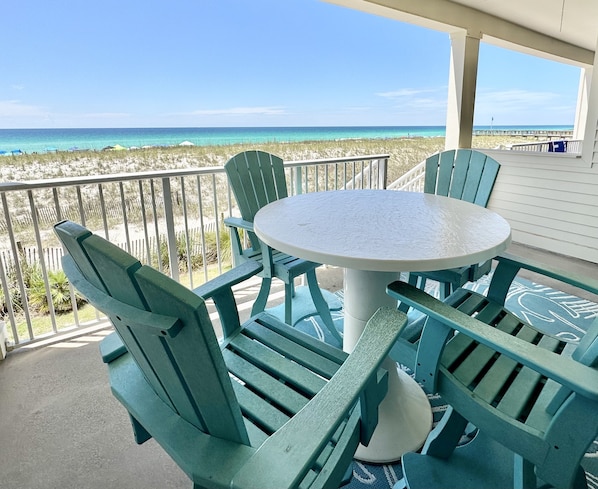 A balcony with a round table and four green chairs, overlooking a beach with clear blue water and a pier in the distance.