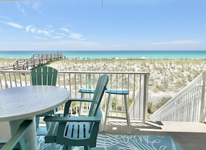 A balcony with a table and chairs overlooking a beach with a wooden walkway and beach huts.