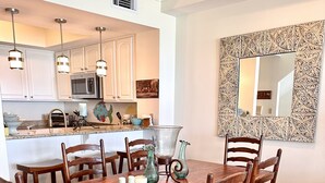 Un coin repas avec une table et des chaises en bois, une cuisine à l’arrière-plan et un miroir décoratif au mur.