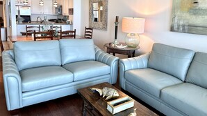 Un salon avec deux canapés en cuir bleu clair, une table basse en bois ornée de motifs de poissons, un coin repas avec des chaises en bois, une cuisine avec des meubles blancs et un tableau abstrait accroché au mur.