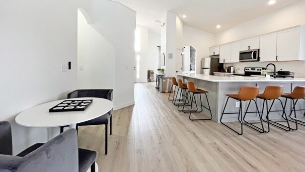 A modern kitchen with white cabinets, a microwave, and a sink. There is a dining area with a round table and chairs.