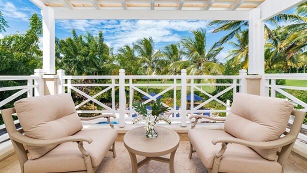 A covered outdoor seating area with two beige chairs and a small round table, surrounded by a white fence with palm trees in the background.
