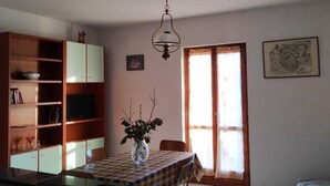 Una habitación con una mesa de comedor, sillas, un televisor en un estante, un jarrón con ramas y una ventana con cortinas.