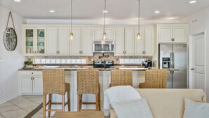 A modern kitchen with white cabinetry, a central island with bar stools, a refrigerator, and a microwave. - Pair-a-dice 3 Bedroom Home by RedAwning (Port Bolivar)