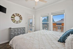 Un dormitorio con una cama grande, un ventilador de techo, un televisor montado en la pared y vistas al balcón exterior.