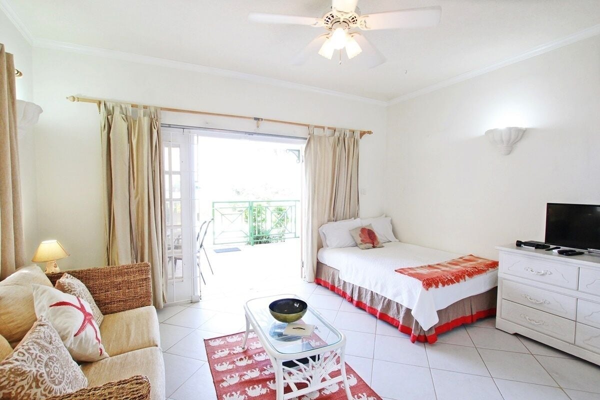 Une chambre à coucher comprenant un lit, une télévision, une commode blanche, une table basse en verre, un tapis à motifs rouges et blancs, et un balcon avec des rideaux.