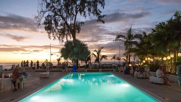 A poolside area with a view of the sunset over the ocean, surrounded by palm trees and people relaxing. - Aqua Bliss 3 Bedroom Home by Bluescape (Holetown)