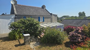 Une maison à deux étages, dotée d’un toit en pente, d’une antenne parabolique et d’un jardin où poussent diverses plantes et fleurs.
