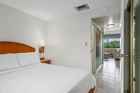 A hotel room with a large bed, a nightstand with a lamp, a door leading to a balcony with a view of greenery.