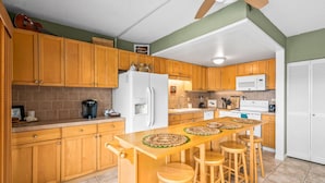 A kitchen with wooden cabinets, a white refrigerator, a microwave, and a bar area with stools. - Big Island Kona Plaza 311 2 Bedroom Condo (Kailua-Kona)