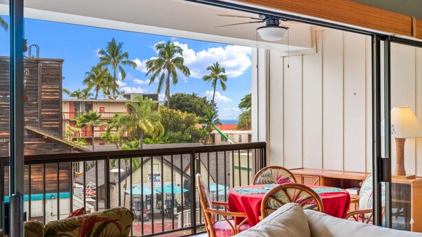 A balcony with a view of palm trees and buildings, a table with a red tablecloth, and wicker chairs. - Big Island Kona Plaza 311 2 Bedroom Condo (Kailua-Kona)