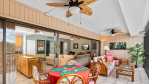 A spacious living area with a large sliding glass door leading to a kitchen, a dining table with chairs, a sofa, and a ceiling fan. - Big Island Kona Plaza 311 2 Bedroom Condo (Kailua-Kona)