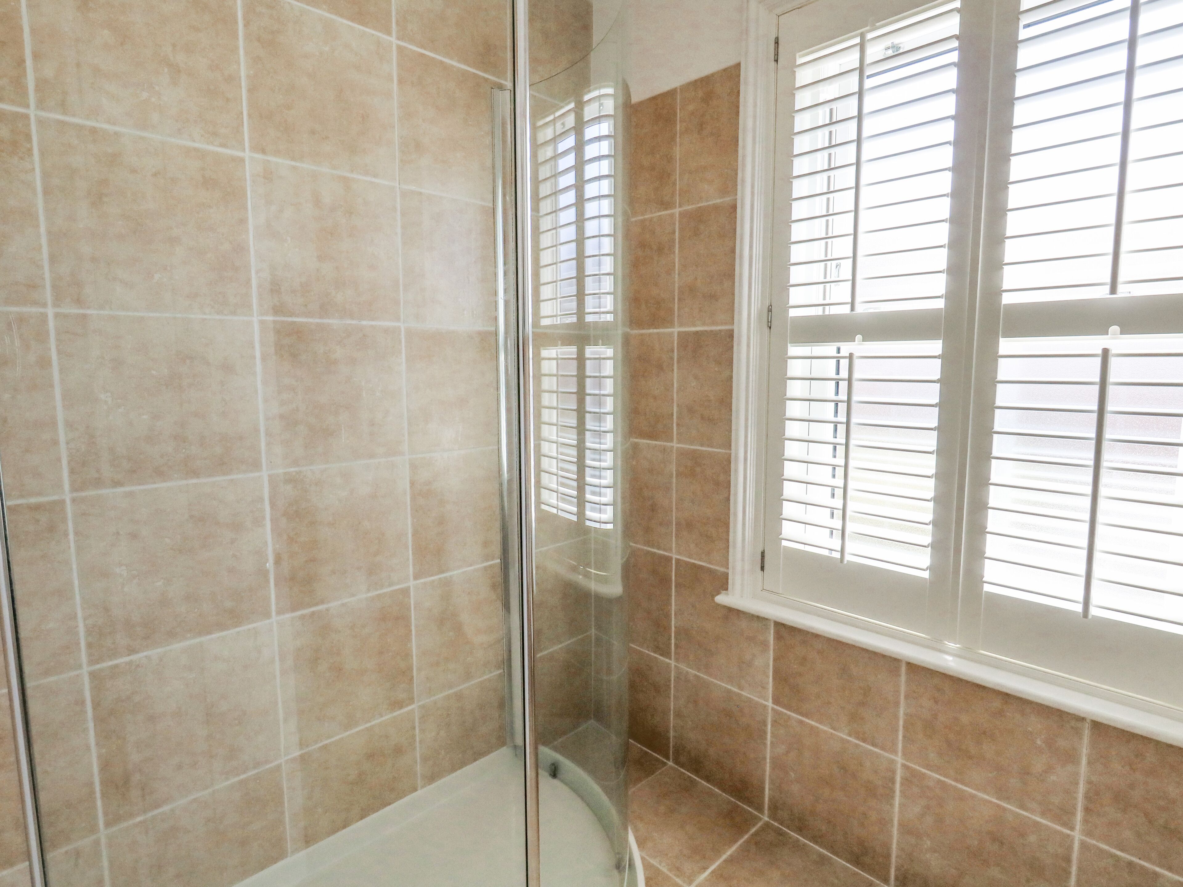 Une salle de bain carrelée de carreaux beiges, dotée d’une douche en verre et d’une fenêtre avec des stores.
