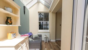 Ett kompakt bostadsutrymme med ett litet kök, en grå soffa, ett litet bord och ett fönster med persienner.