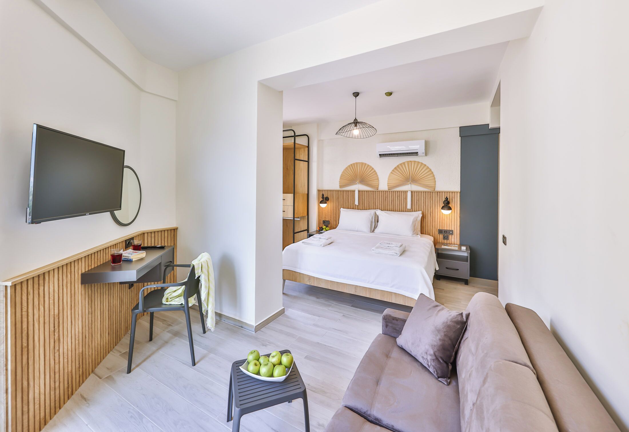 A modern hotel room with a bed, a sofa, a desk with a flat-screen TV, a small table with apples, and a wooden accent wall.