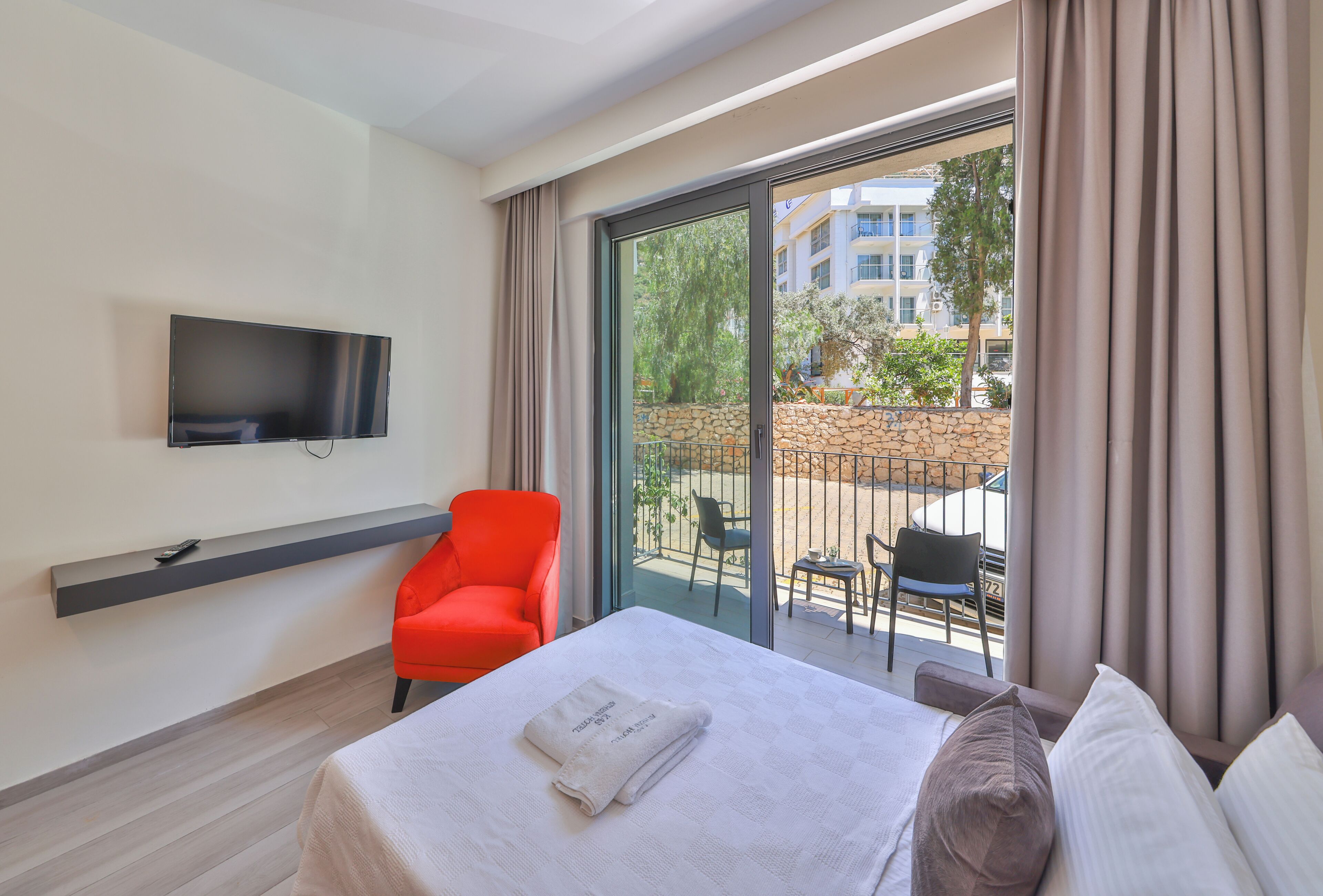 A hotel room with a bed, a red chair, a TV mounted on the wall, and a balcony with outdoor seating.