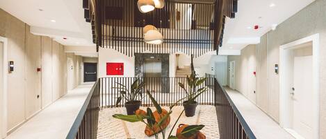 A modern hotel lobby with a central staircase, potted plants, and a red emergency box.