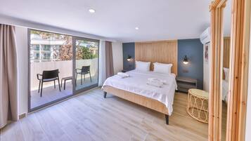 A hotel room with a large bed, a wooden floor, a balcony with chairs, and a view of the city.