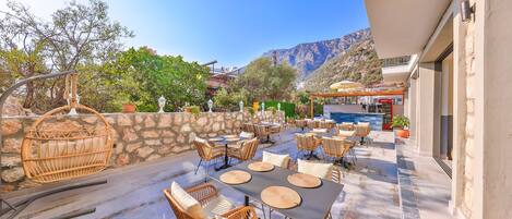 An outdoor patio with wicker furniture and tables, stone walls, and a mountain backdrop.