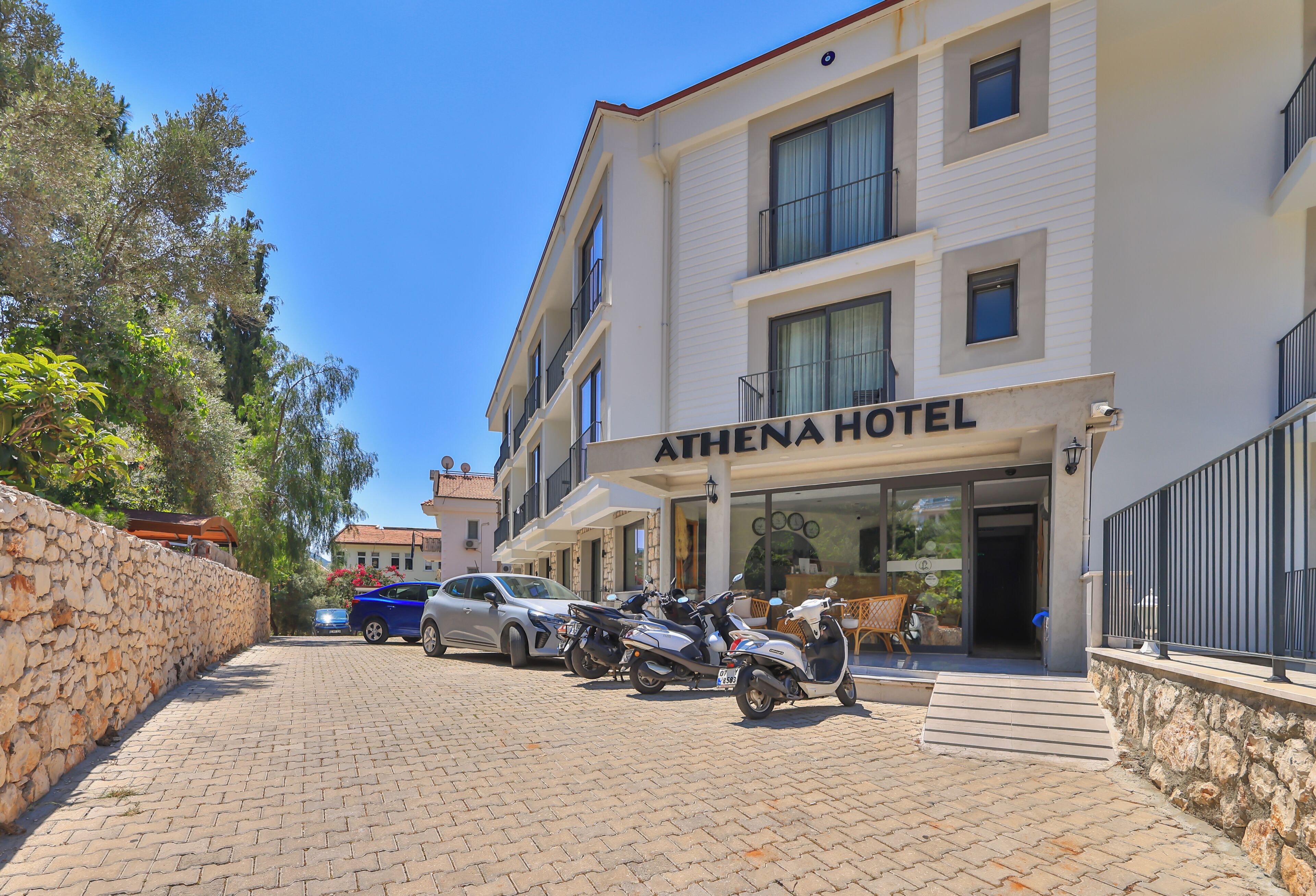 A hotel named 'ATHENA HOTEL' with a paved entrance, parked cars, and a stone wall on the left.