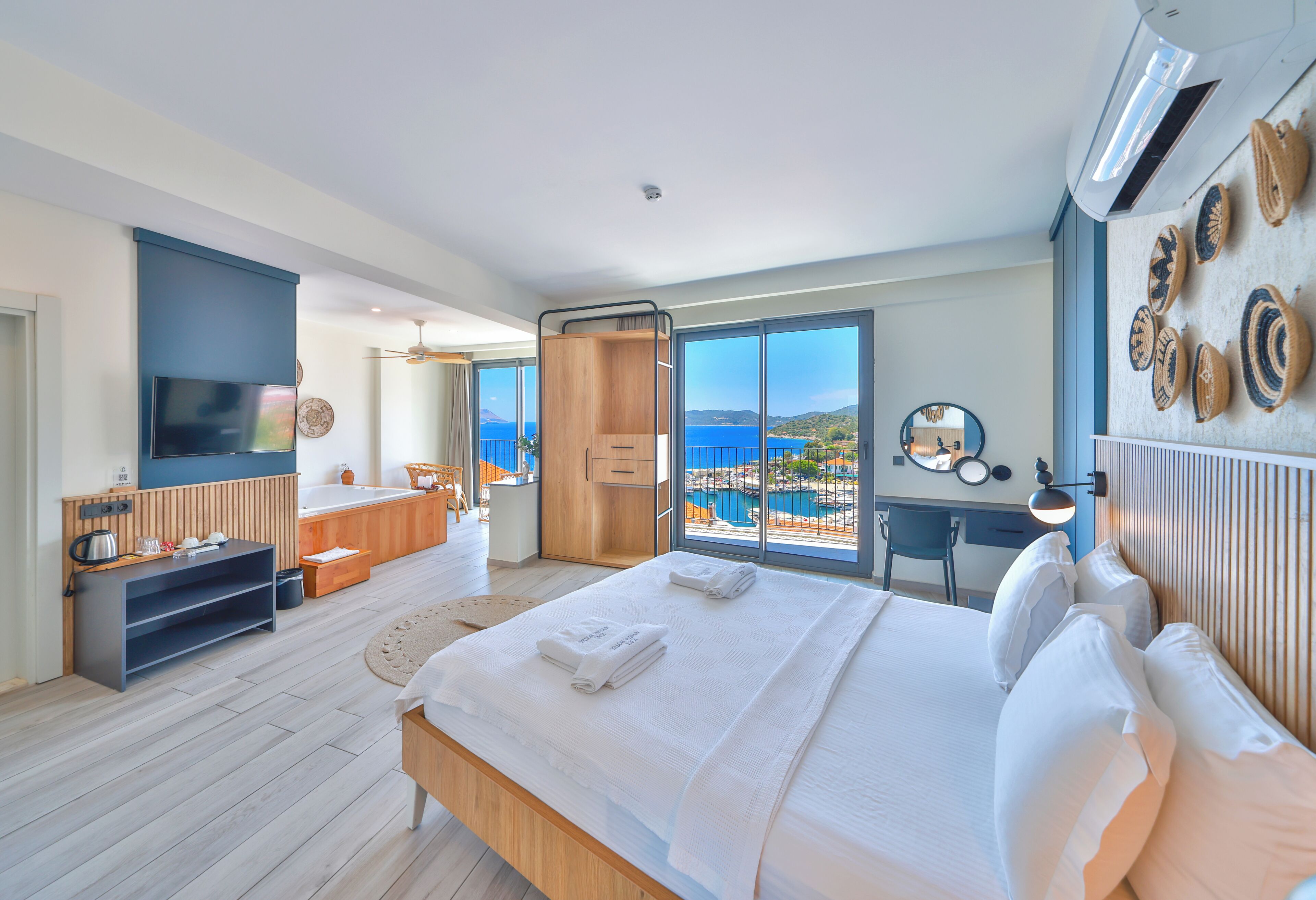 A modern hotel room with a large bed, a TV, a desk, and a view of the sea.