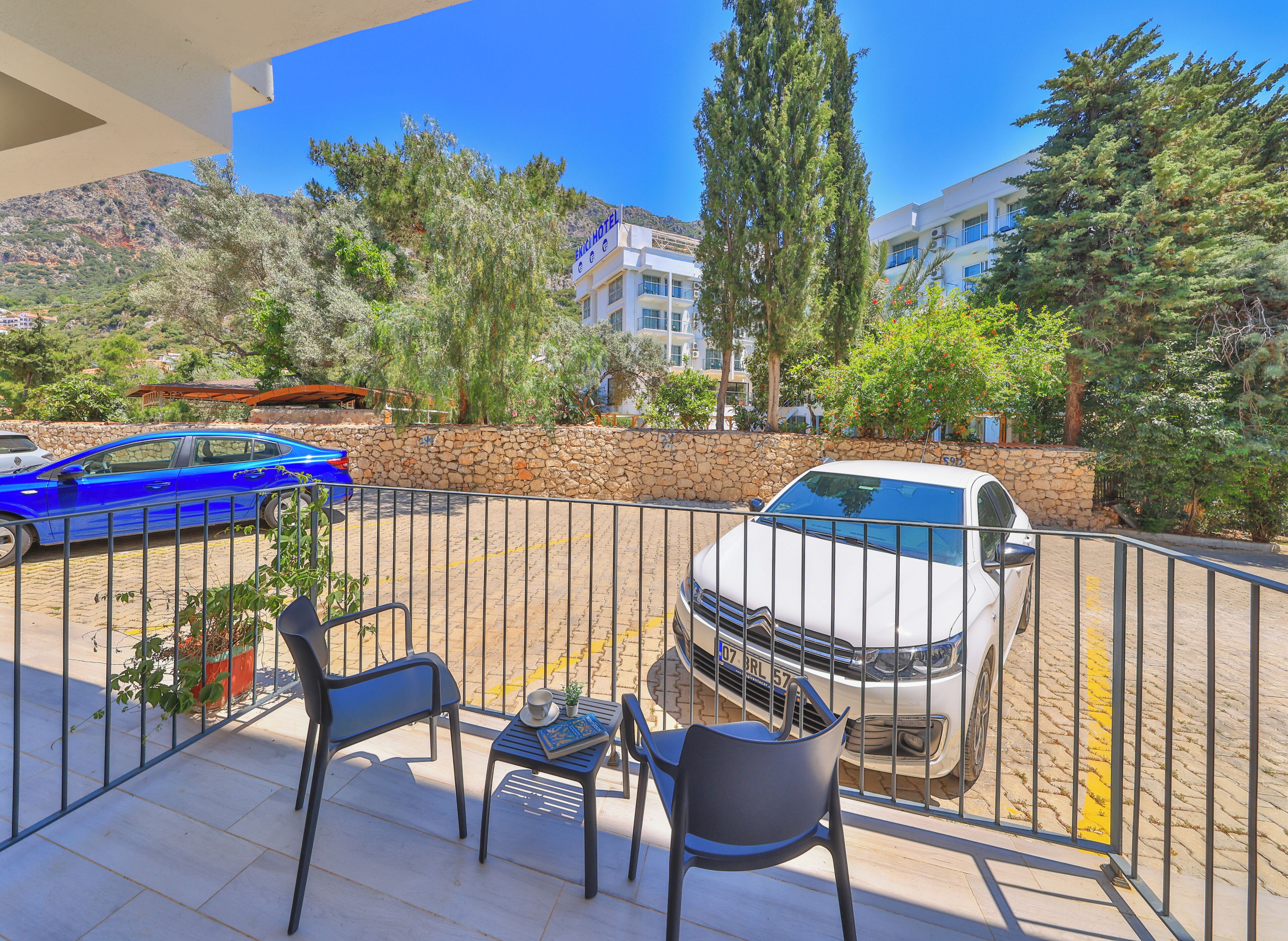 Un balcón con un coche blanco estacionado al fondo, un muro de piedra y vistas a un hotel y montañas.