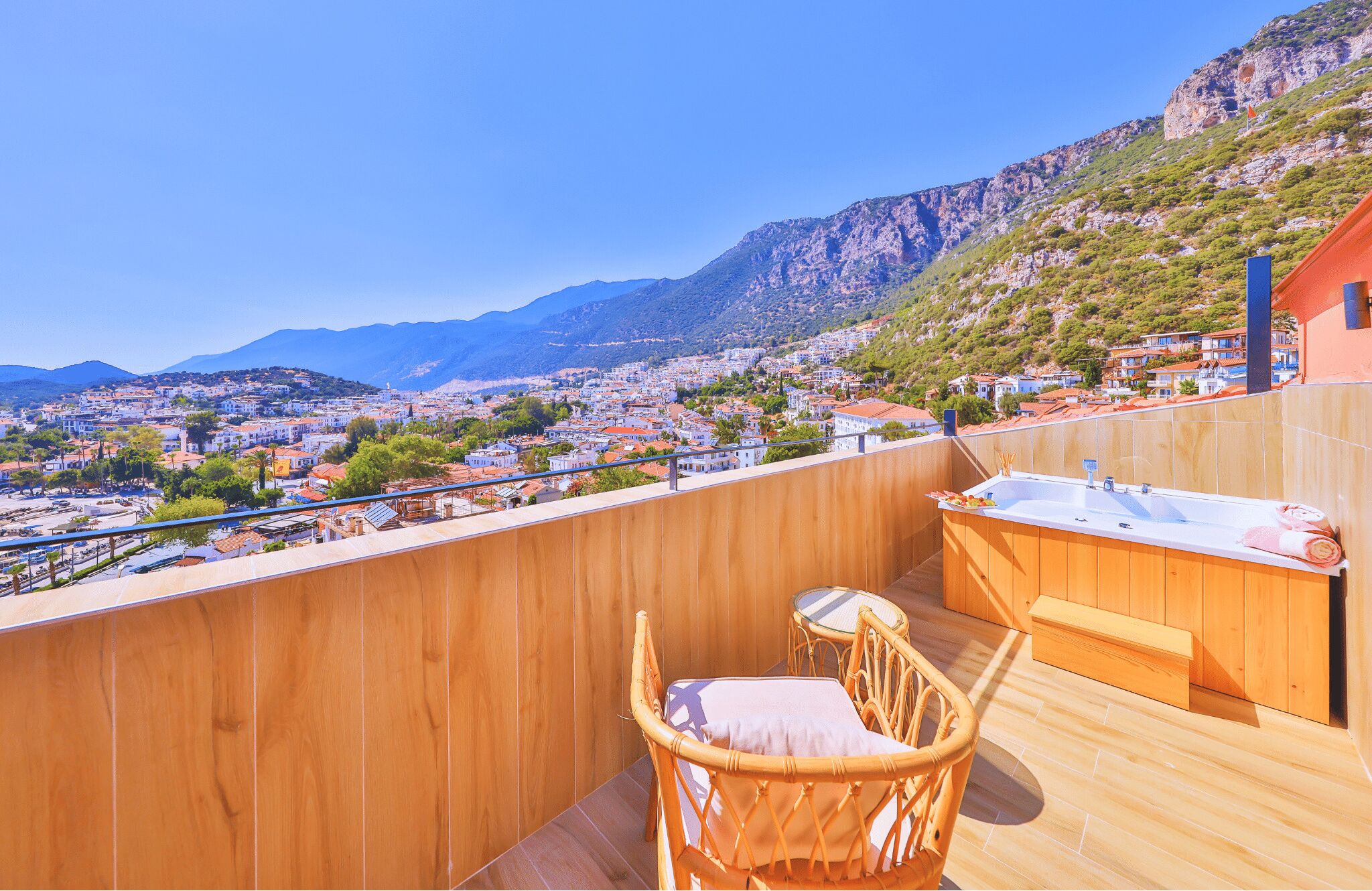 Terraza de madera con una silla de mimbre y una pequeña mesa, con vistas a un pueblo y montañas al fondo.
