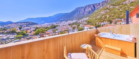 Terraza de madera con una silla de mimbre y una pequeña mesa, con vistas a un pueblo y montañas al fondo.