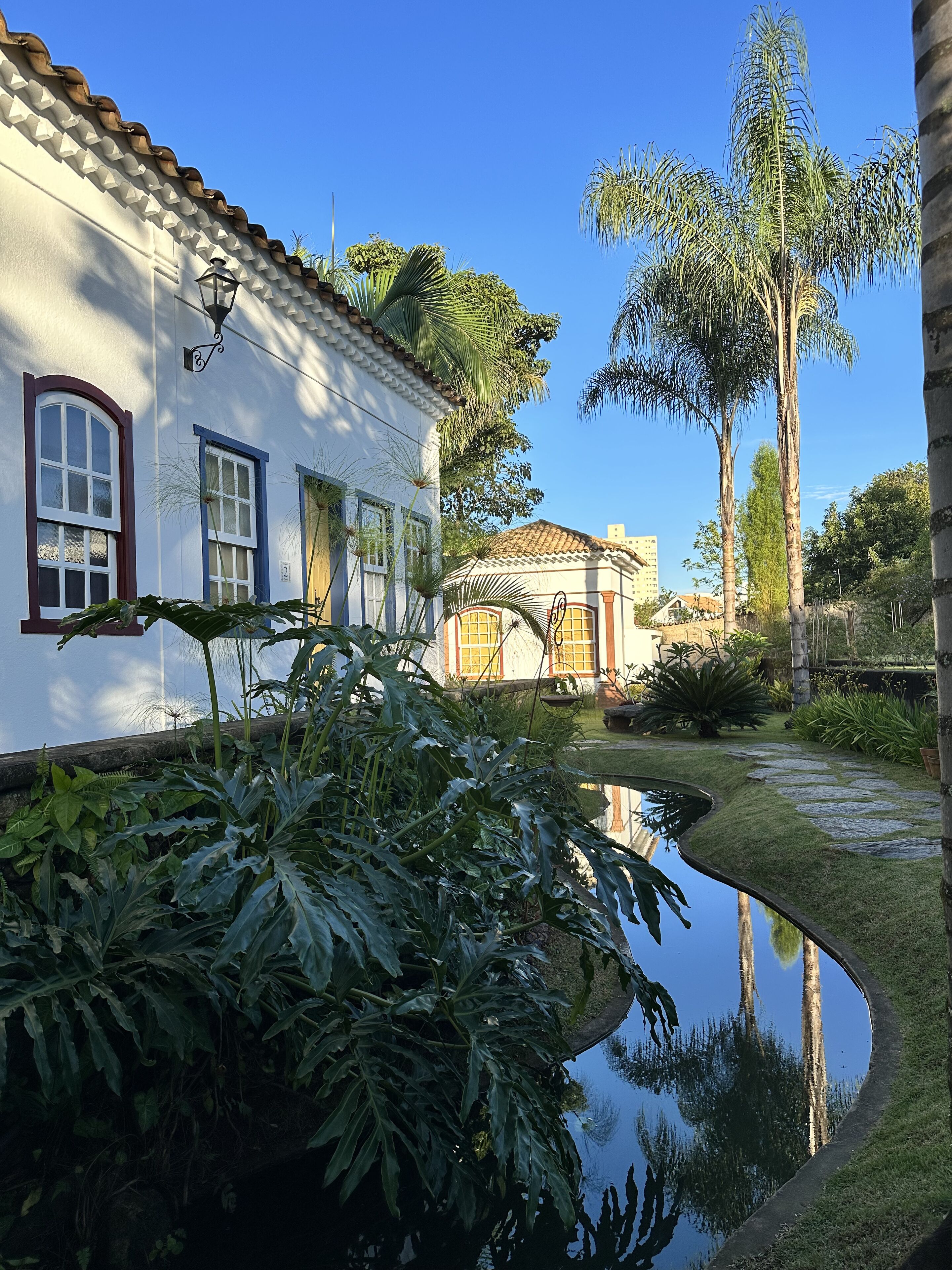Um edifício branco com telhado ladrilhado, um lago refletindo a paisagem e vegetação exuberante.