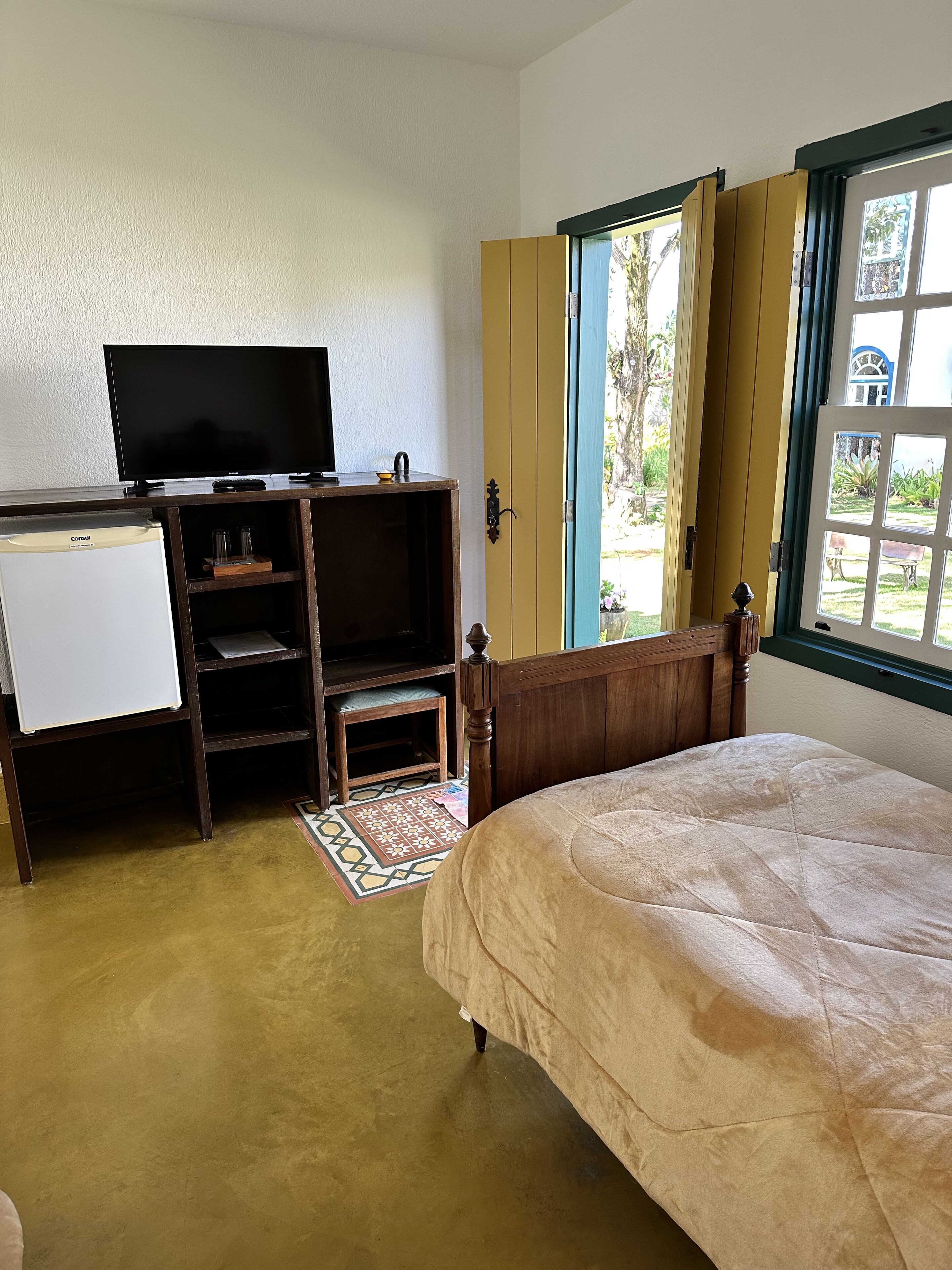 Un dormitorio con cama, televisor sobre un mueble de madera, refrigerador pequeño y ventana con cortinas.