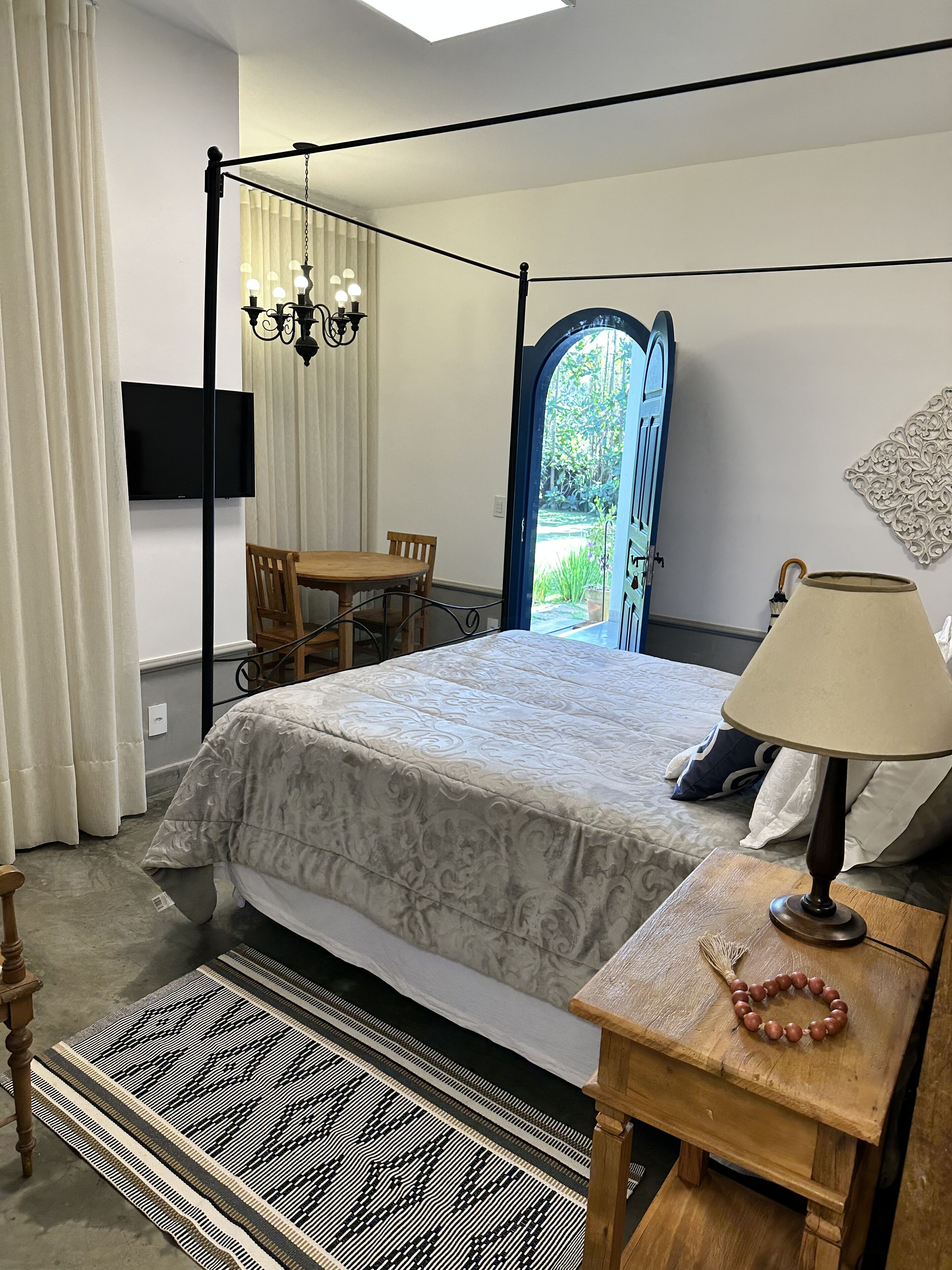 Un dormitorio con cama con dosel, una mesita de noche de madera con una lámpara, un televisor montado en la pared y vistas al jardín a través de una puerta.