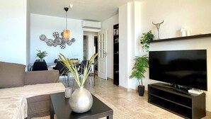 Ein modernes Wohnzimmer mit einer Couch, einem Couchtisch mit einer Vase, einem Flachbildfernseher auf einem Schrank und einem Essbereich im Hintergrund.