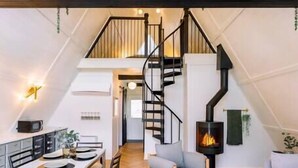 A loft-style room with a wooden floor, a dining area with chairs and a table, a sofa, a staircase, and a fireplace. - Secluded A-Frame Hocking Hills: Hot Tub, Fire Pit (South Bloomingville)