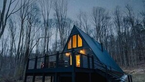 A cabin with a sloped roof, a balcony, and a lit interior. - Secluded A-Frame Hocking Hills: Hot Tub, Fire Pit (South Bloomingville)
