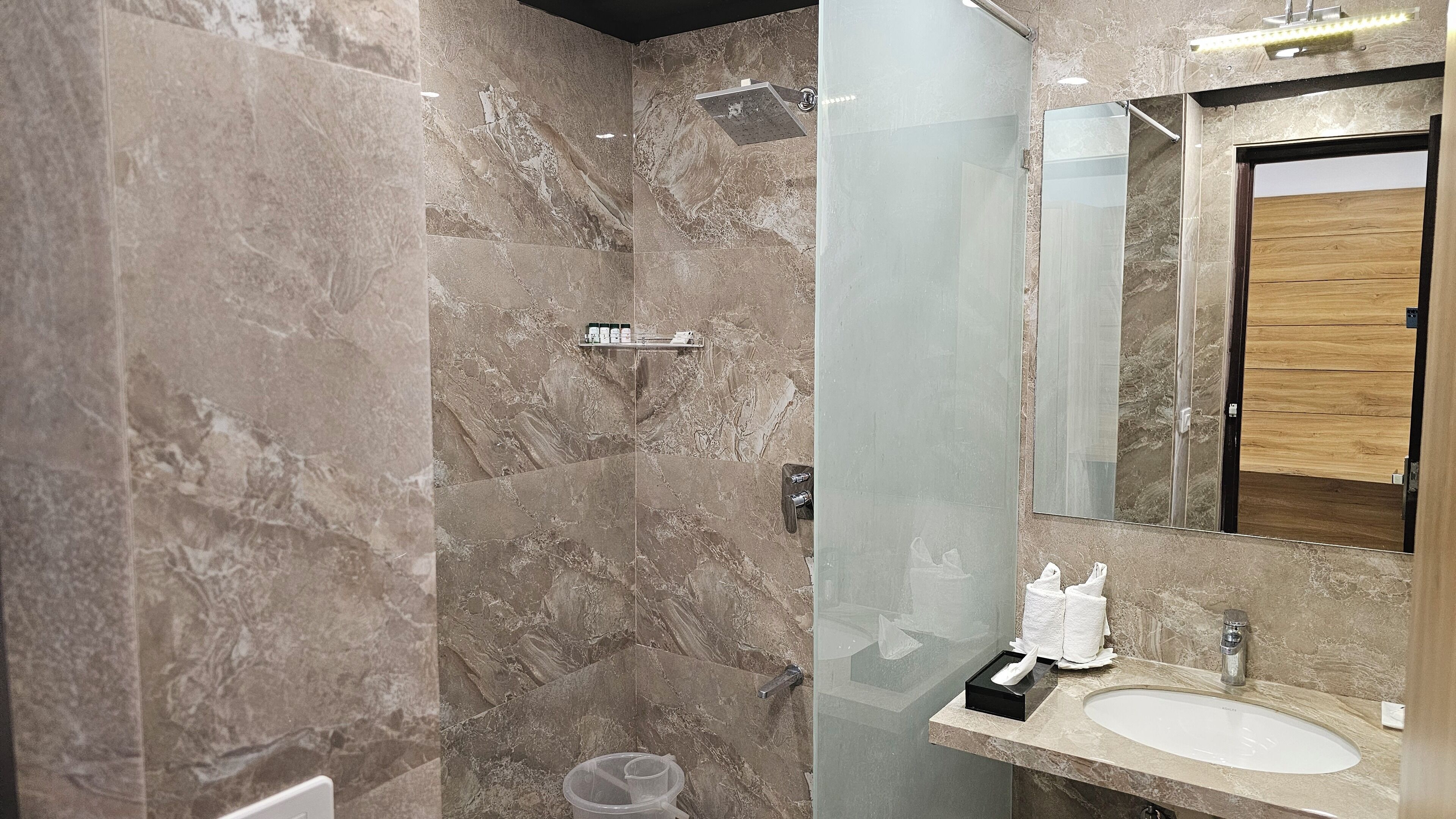 A modern bathroom with a glass shower enclosure, a marble wall, a wooden vanity, and a large mirror.