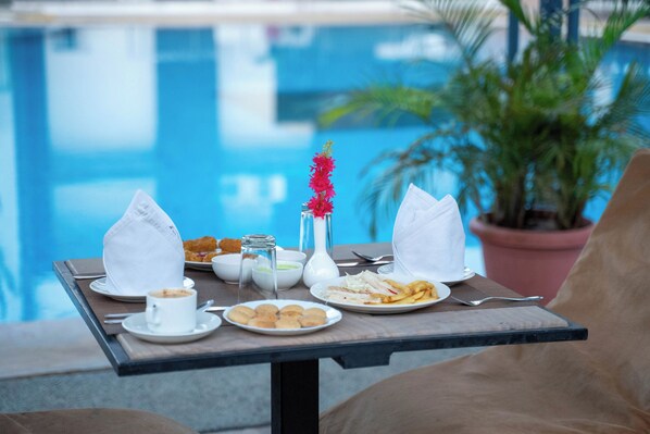 Una mesa con alimentos para el desayuno junto a una piscina, incluyendo platos de comida, una taza y un florero con flores.
