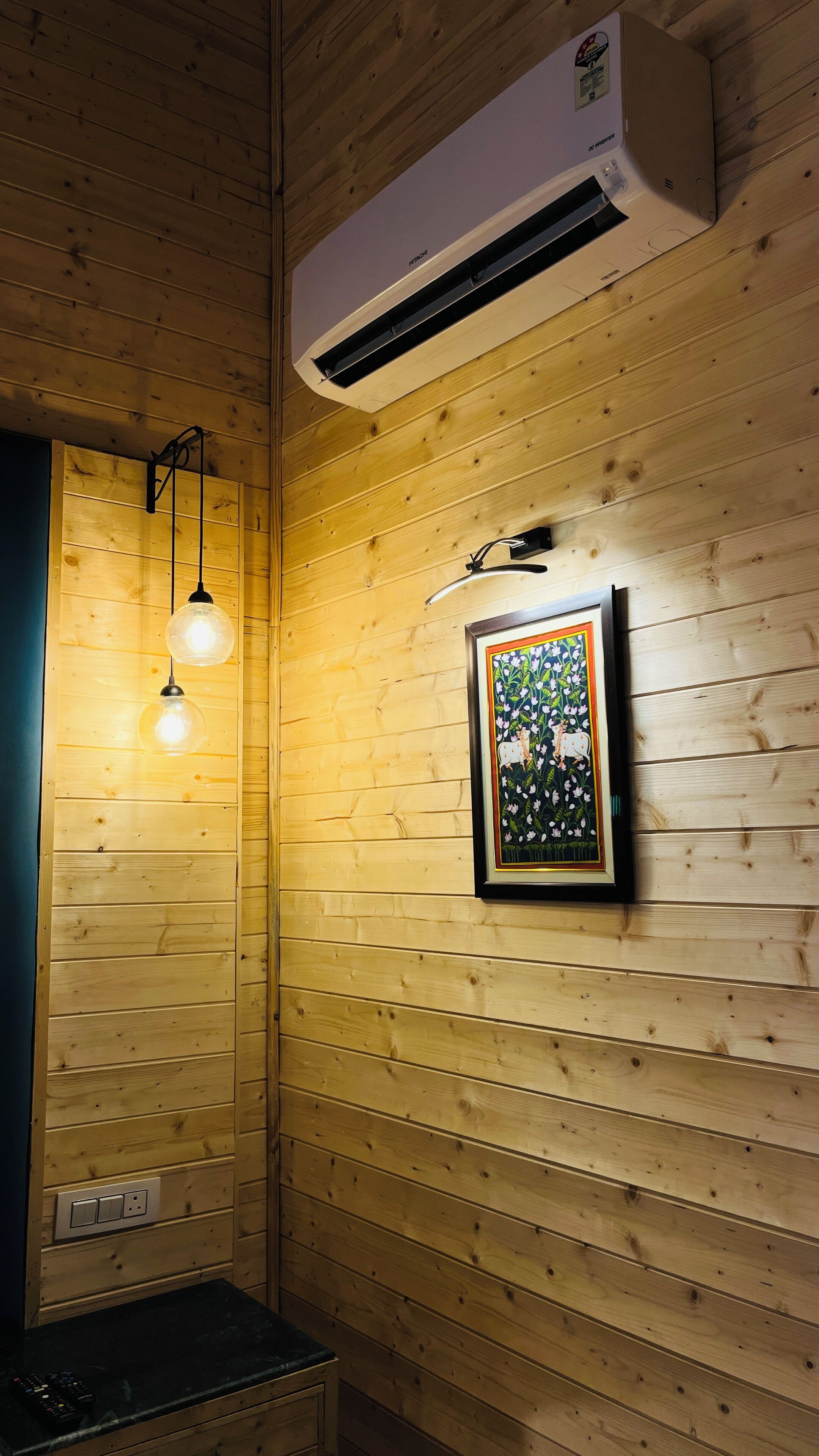 A wooden cabin interior with a mounted air conditioner, a framed picture on the wall, and two hanging light fixtures.