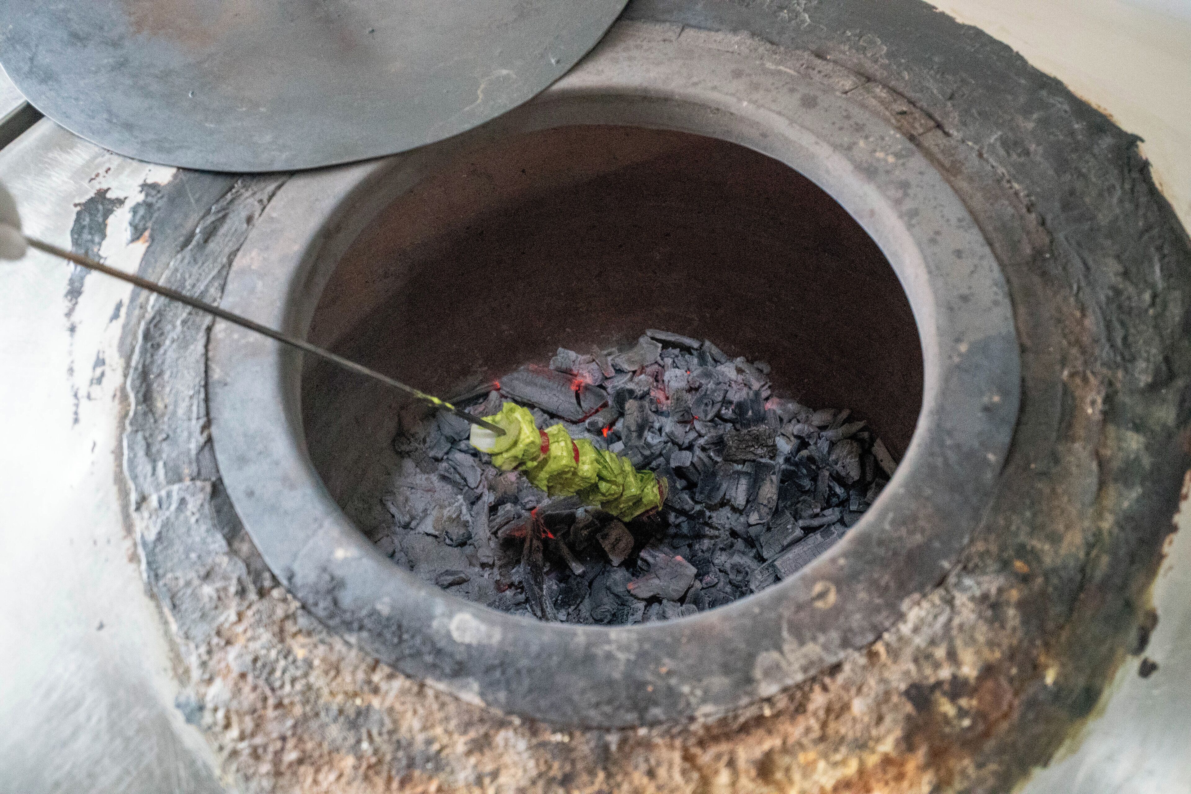 A traditional cooking method with a large stone pot containing charcoal and a green leafy vegetable being cooked over an open flame.