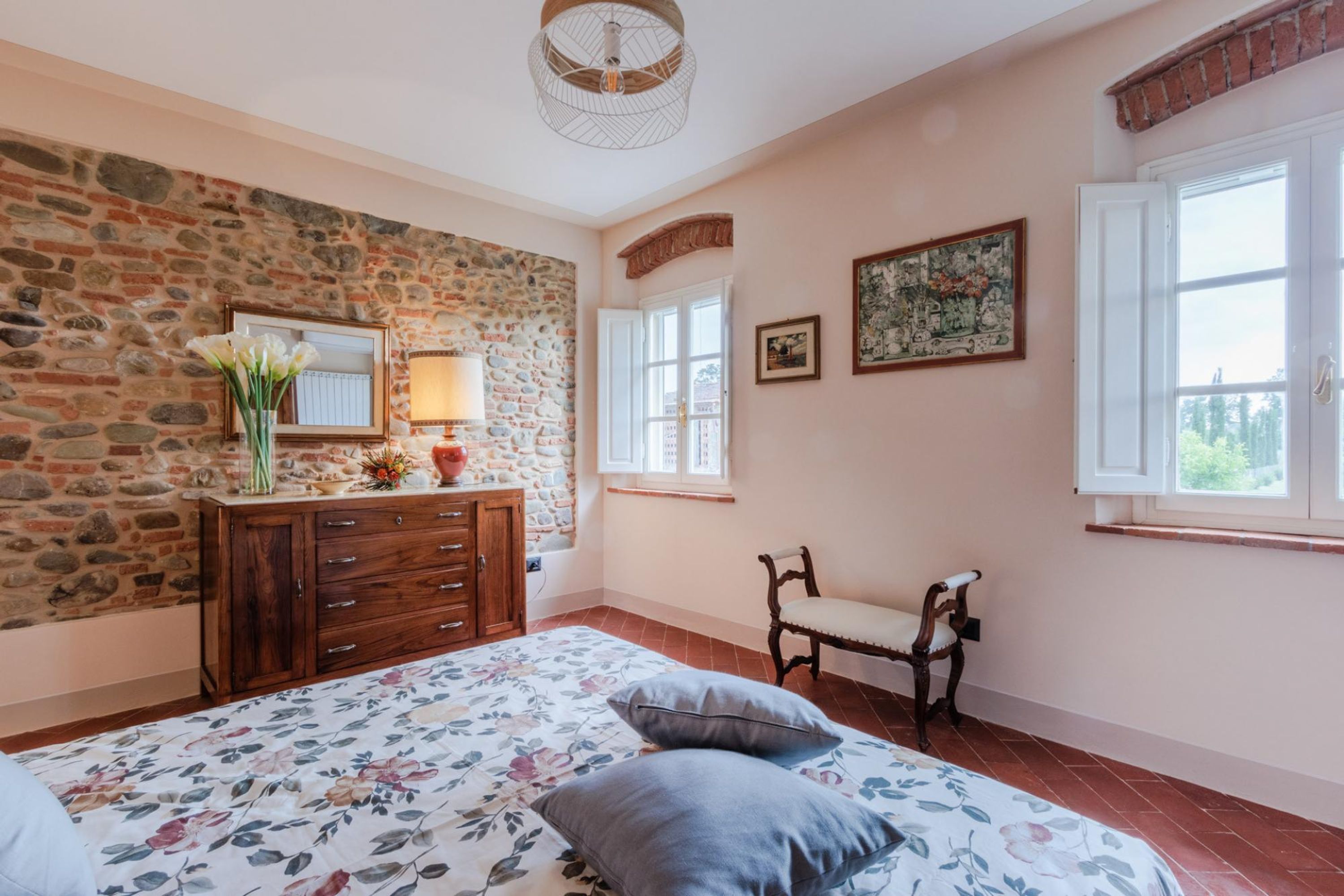 Een slaapkamer met een stenen muur, een houten dressoir, een bloemenprint beddeken en een stoel bij het raam.