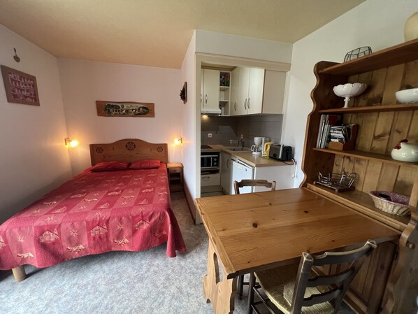 A bedroom with a bed, bedside tables, and a wooden dining table with chairs. There is a kitchen area with cabinets and a sink.