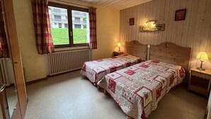 Un dormitorio con dos camas, cabeceros de madera, una ventana con cortinas, un radiador y una mesita de noche con una lámpara.
