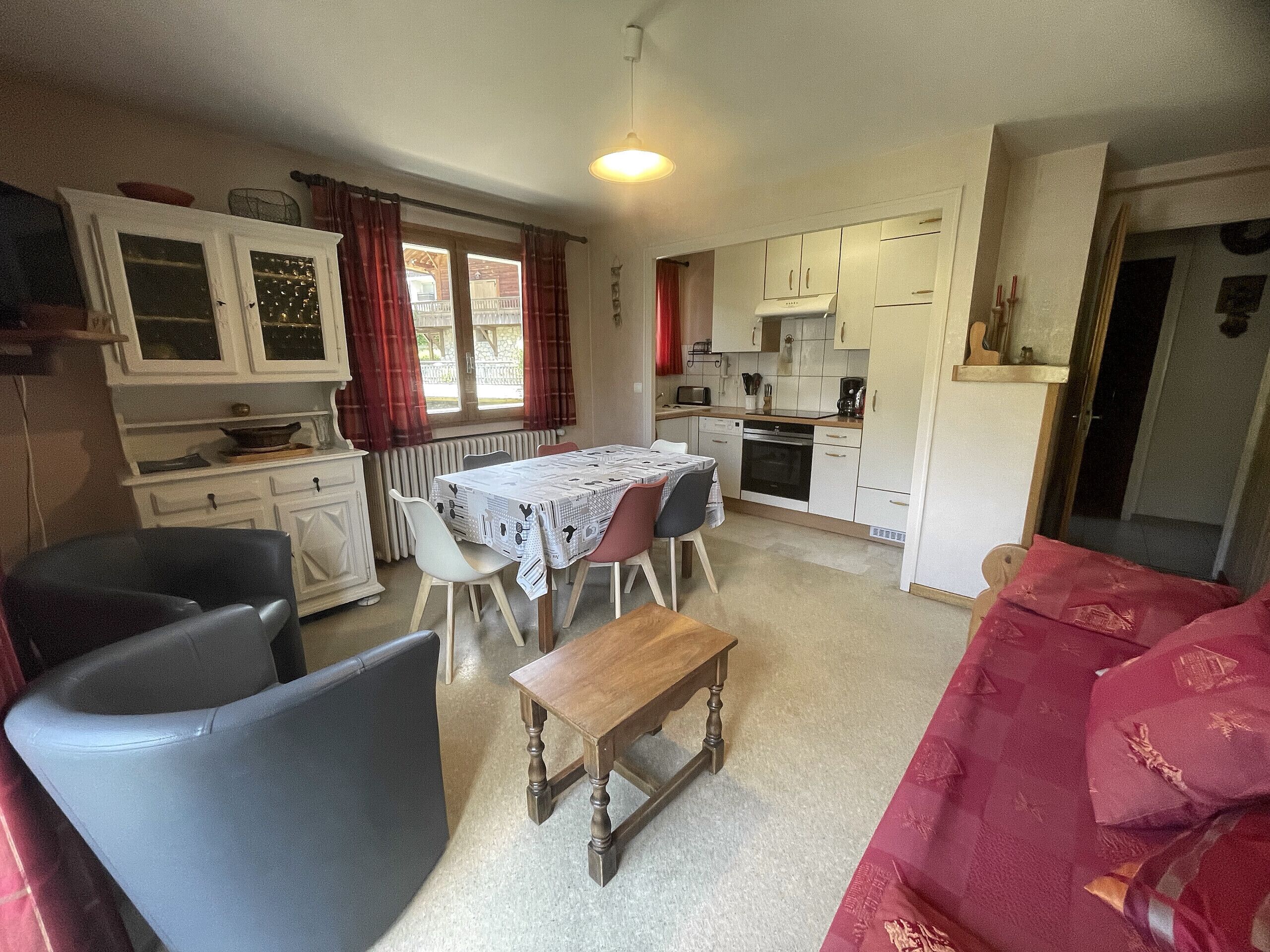 A compact kitchen with white cabinets, a dining table with chairs, a bench, and a red sofa.