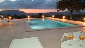 Piscina rectangular al aire libre con vistas a montañas al atardecer. Hay sillones de descanso y mesas con copas de vino.