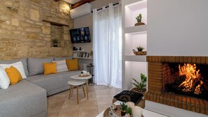 Una sala de estar con pared de piedra, un sofá gris con cojines amarillos, una mesita, un televisor montado en la pared, una chimenea encendida y una cortina blanca.