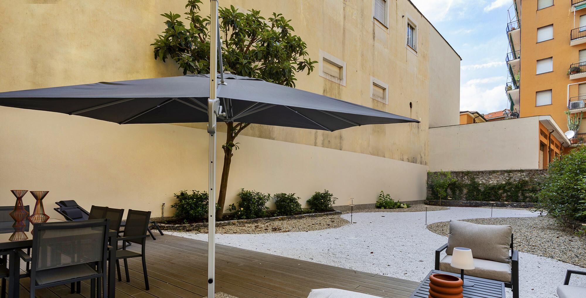An outdoor patio area with a table and chairs, a large umbrella, and a small garden with plants and a tree.