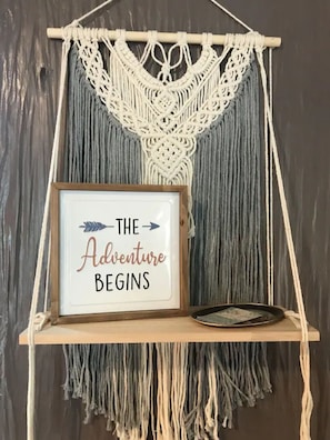 A macrame wall hanging with a framed sign that reads "THE Adventure BEGINS".