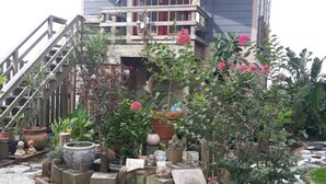 Ein zweistöckiges Haus mit Balkon, umgeben von einem Garten mit verschiedenen Kübelpflanzen und einem kleinen Bach.